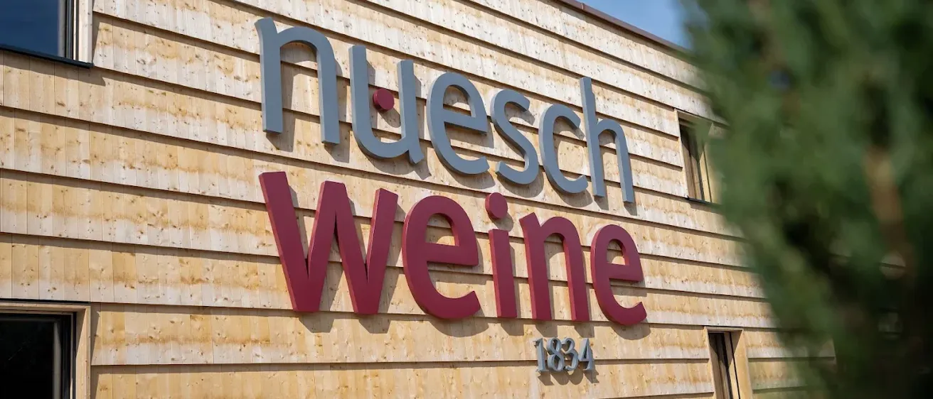 Die Vorderseite eines Holzgebäudes zeigt ein Schild mit der Aufschrift 'nuesch weine' und darunter ein rotes '1834'. Das Schild ist an einer Holzfassade befestigt.