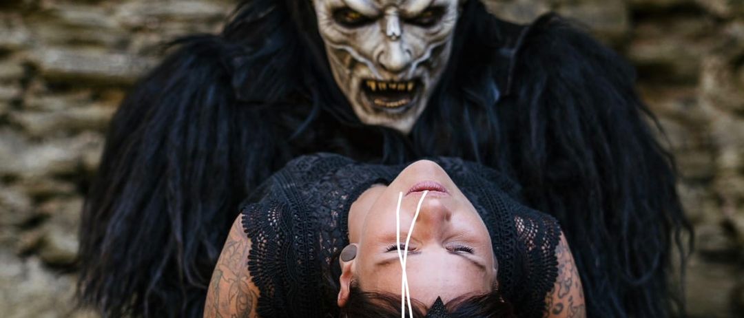Eine tätowierte Frau liegt auf dem Rücken, während eine gehörnte Dämonenmaske über ihr schwebt. Beide sind in dunkler Kleidung gekleidet, die Frau trägt ein schwarzes Spitzenoberteil.