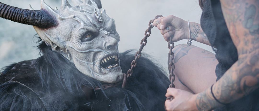 Eine Frau mit Tattoos sitzt auf einem schwarz behaarten Geschöpf und hält eine Kette um seinen Hals. Das Geschöpf hat Hörner und eine weiße Maske. Die Szene ist neblig.