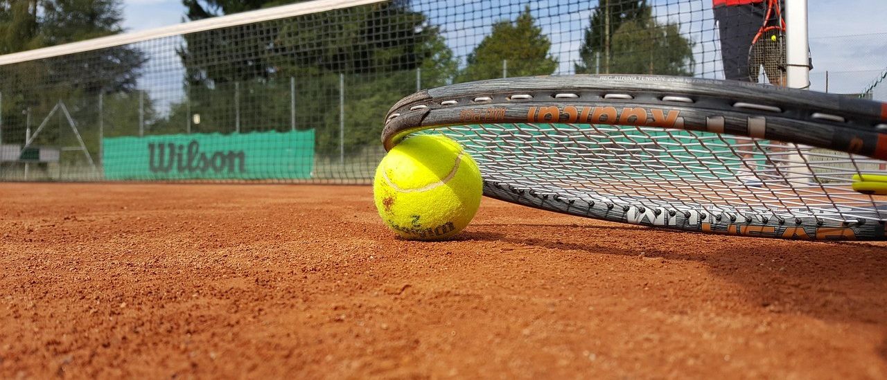 Ein Tennisball liegt auf einem Tennisplatz, neben einem Tennisschläger. Der Platz hat braunen Boden, ein Netz im Hintergrund und ein Banner mit der Aufschrift Wilson.