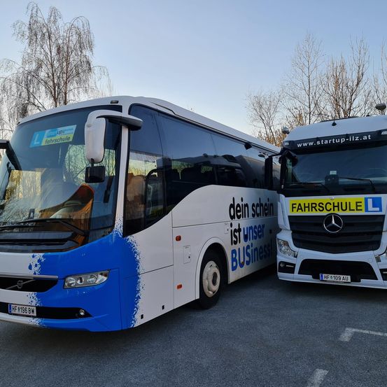 Zwei Busse, einer mit einem blau-weißen Farbschema und der andere weiß, sind auf einem Parkplatz geparkt. Der blau-weiße Bus hat die Aufschrift 'Dein Schein ist unser Business!' an der Seite.