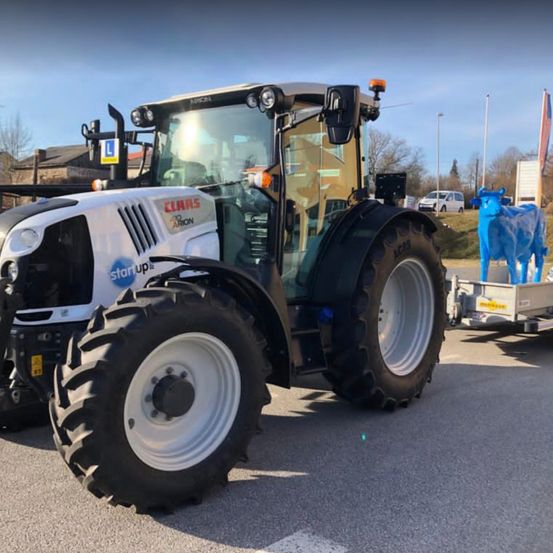Ein weißer Traktor mit 'CLAAS' und 'start up' Branding steht auf einer Straße, in der Nähe einer blauen Kuhstatue.