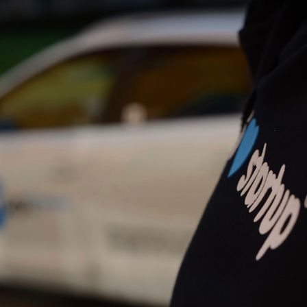 Ein Nahaufnahme eines schwarzen Shirts mit dem Wort 'storop' in Weiß auf der linken Seite. Dahinter ist ein verschwommenes weißes Auto zu sehen.