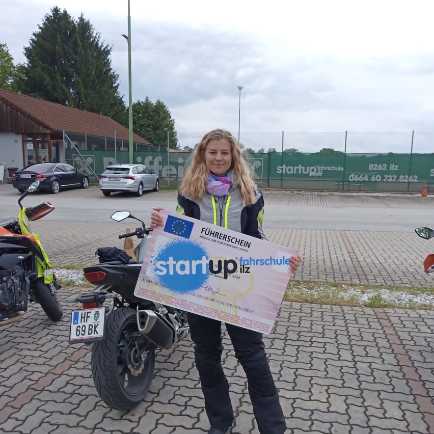 Eine Frau hält ein Zertifikat mit der Aufschrift Start up Fahrschule vor einem Motorrad und einigen geparkten Autos.