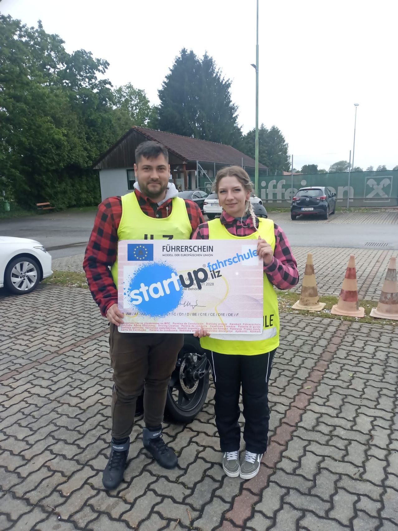 Zwei Personen in gelben Warnwesten stehen draußen und halten ein Zertifikat mit der Aufschrift 'Startup ilz'. Sie stehen neben einem geparkten Motorrad, mit einem Gebäude und geparkten Autos im Hintergrund.