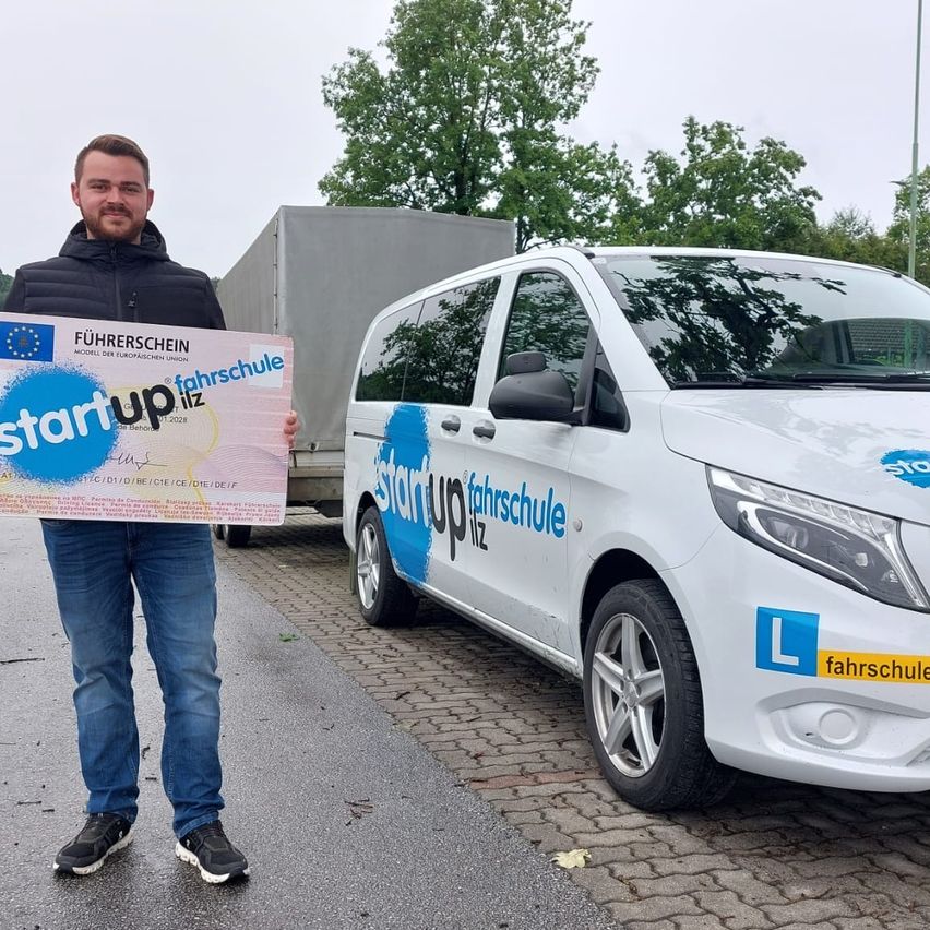 Ein Mann hält ein Schild mit Logo und Text vor einem weißen Van mit blau-weißen Aufklebern auf einer gepflasterten Straße.