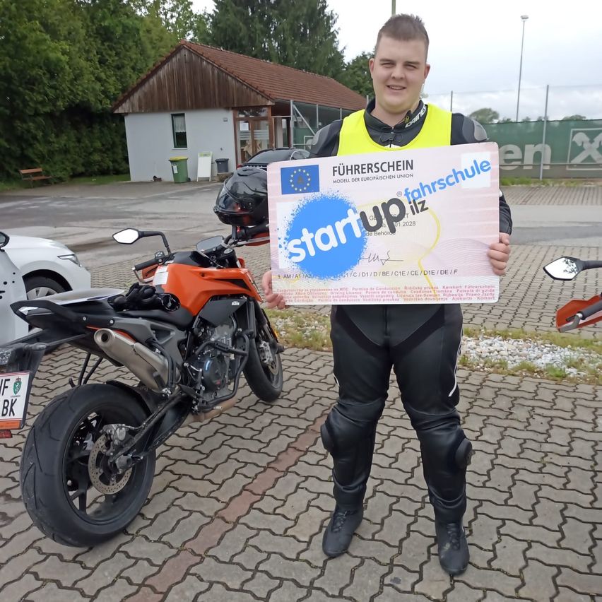 Ein Mann steht neben einem orangefarbenen Motorrad und hält ein Zertifikat in der Hand. Er trägt einen gelben Weste und schwarze Lederhosen.