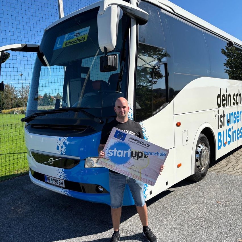 Ein Mann posiert mit einem Zertifikat vor einem Bus mit der Aufschrift 'Start up' und 'Fahrschule' an einem sonnigen Tag.