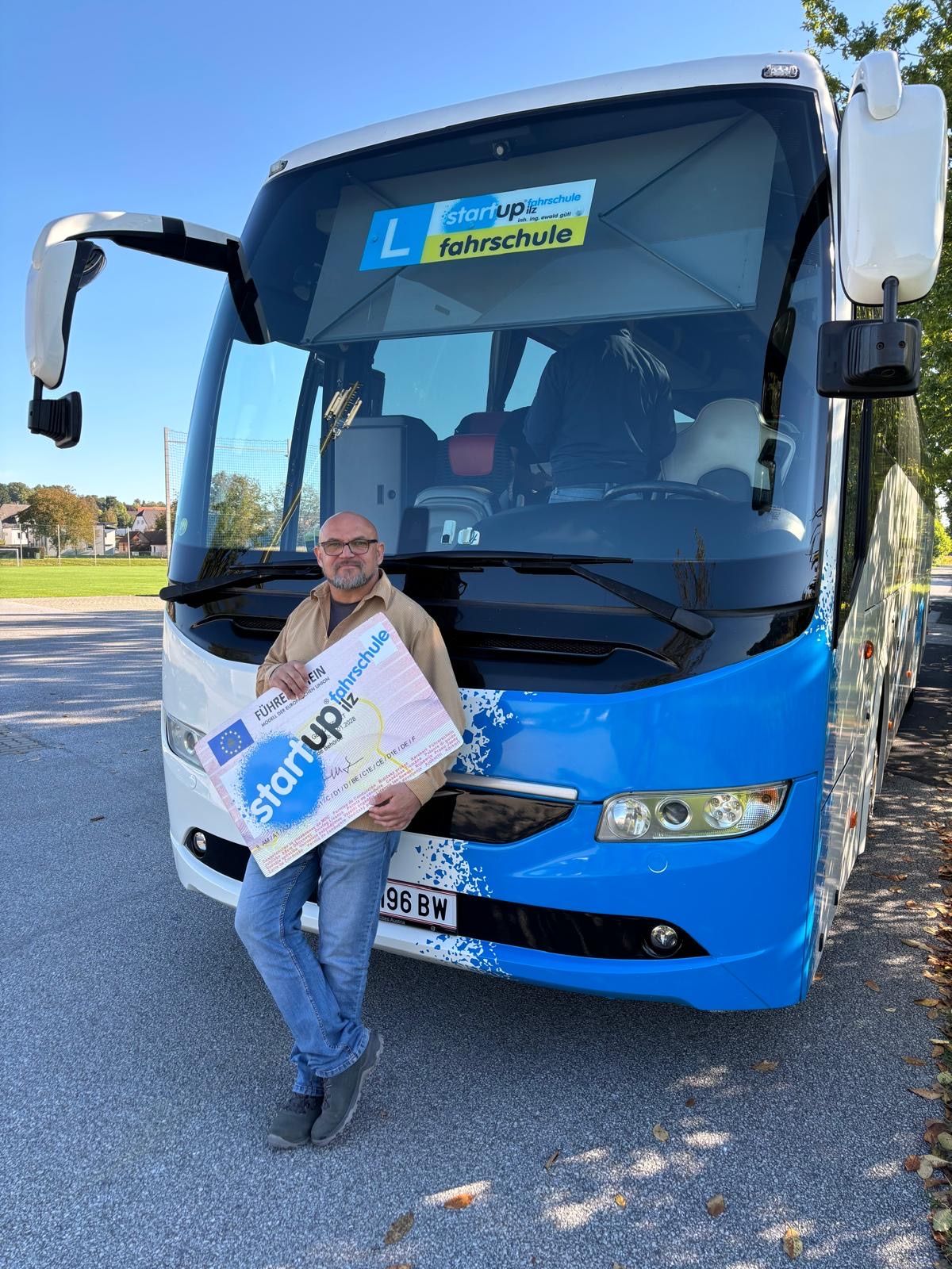 Ein Mann posiert mit einem Zertifikat vor einem Bus und hält ein großes Schild mit der Aufschrift 'Start Up'. Der Bus ist blau und weiß, mit dem Kennzeichen '96 BW'. Hinter dem Bus befindet sich eine Rasenfläche mit Bäumen.