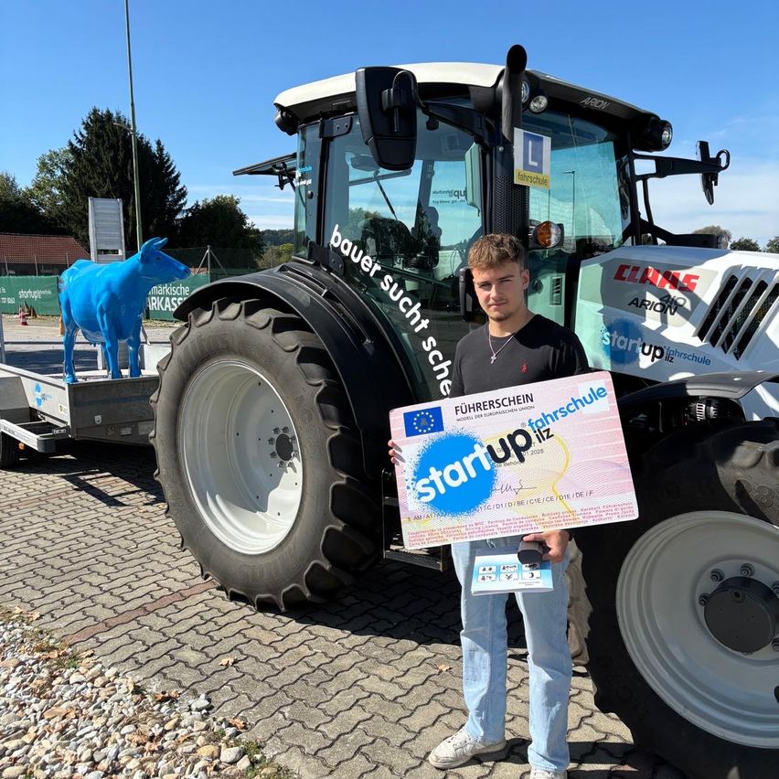 Ein Mann hält ein Zertifikat vor einem Traktor mit einer blauen Kuhstatue im Hintergrund.