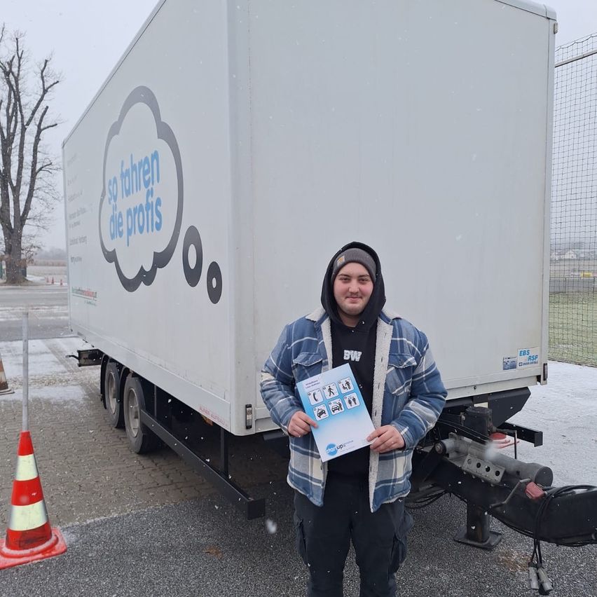 Ein Mann in einer Kapuzenjacke steht vor einem weißen LKW mit einem deutschen Satz und hält ein blaues Heft mit einem Bild von Menschen.