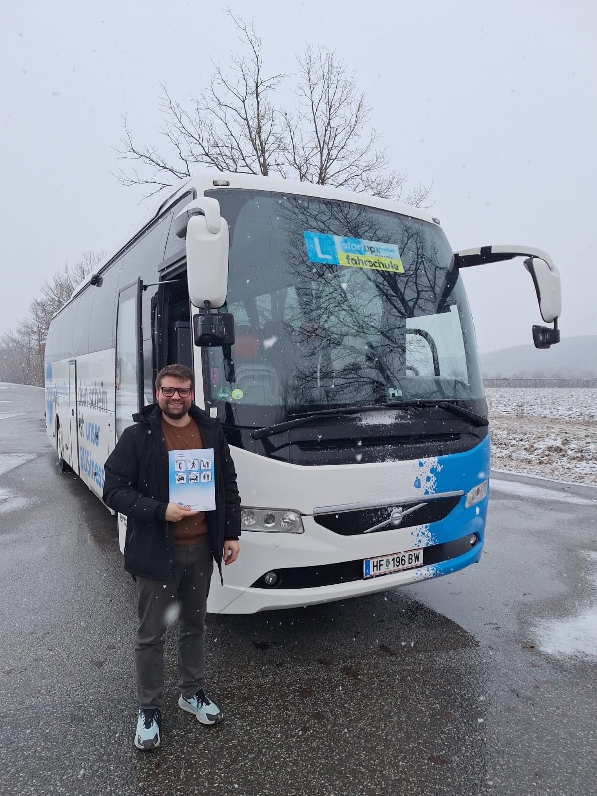 Ein Mann in einem Wintermantel steht neben einem blau-weißen Bus mit einem Fahrschulschild. Der Bus ist auf einer verschneiten Straße geparkt.