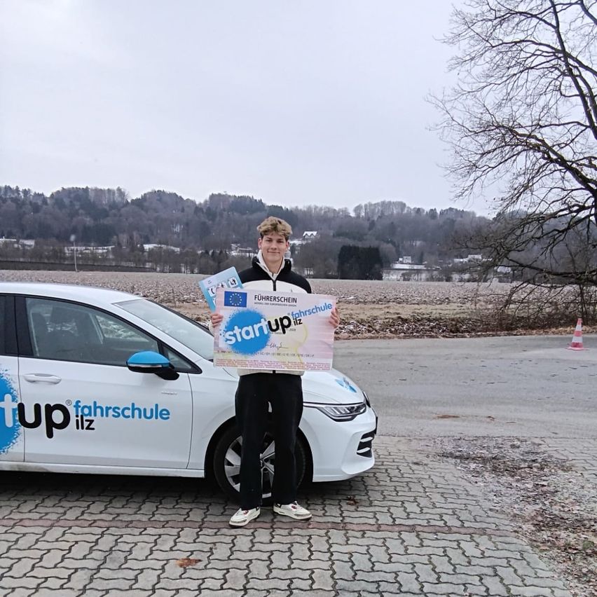 Ein junger Mann steht vor einem weißen Auto mit einem blauen Schild, auf dem 'Startup ilz' steht. Er hält ein Zertifikat in der Hand und lächelt.