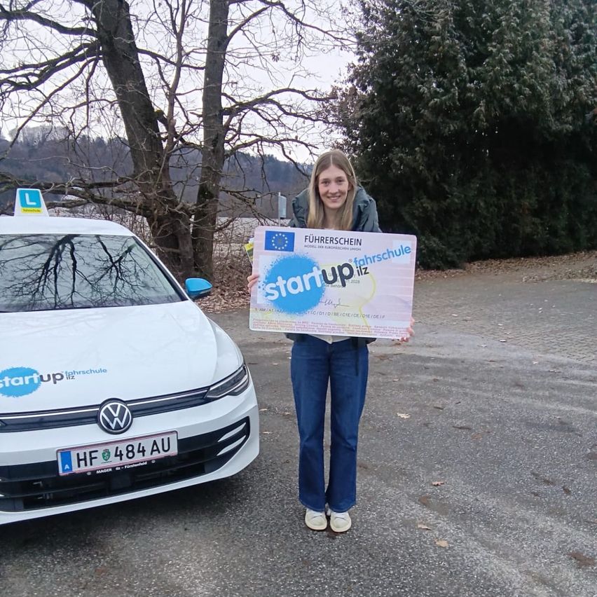 Eine Frau hält ein Zertifikat in der Hand und steht neben einem weißen Auto mit dem Kennzeichen HF 484 AU. Auf dem Zertifikat steht 'Startup liz'. Das Auto hat ein 'StartUp liz' Logo auf der Vorderseite.