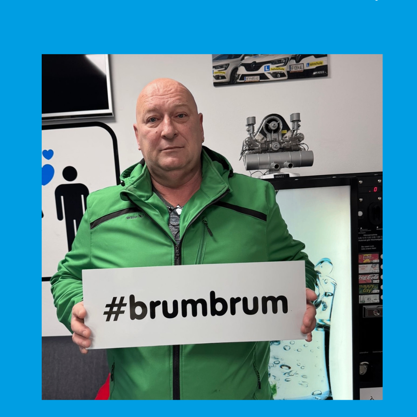 An older man in a green jacket holds a sign that reads #brumbrum. Behind him is a vending machine with sodas.