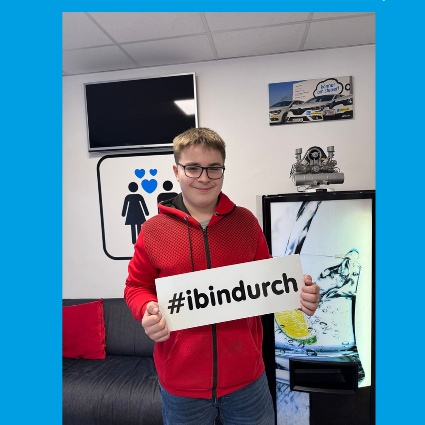 A man in a red jacket holding a sign that says #ibinddurch stands in a room with a sofa, pillows, a television, and a water dispenser.