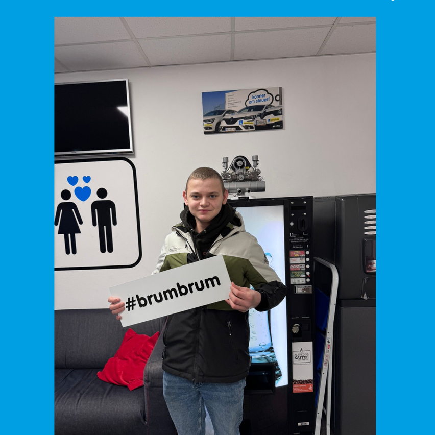 A young man stands in front of a vending machine, holding a sign with #brumbrum on it. Behind him is a wall with a picture of two women and a TV.