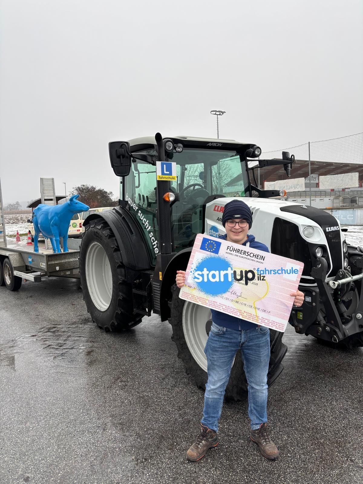 Eine Person hält ein Zertifikat vor einem Traktor mit einer blauen Kuhstatue auf dem Anhänger, unter einem bewölkten Himmel.