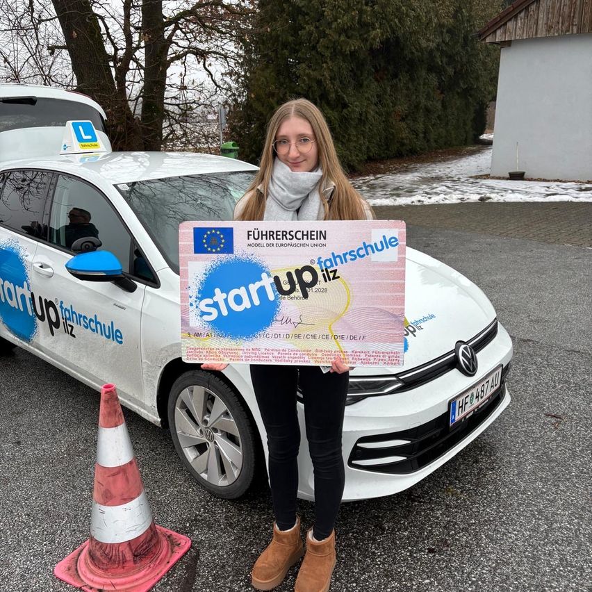 Eine Frau steht vor einem weißen Auto mit der Aufschrift 'Startup Fahrschule'. Sie hält ein Zertifikat mit 'Startup Fahrschule' und einer Karte in der Hand. Das Auto hat ein Taxischild und Schnee auf dem Boden.