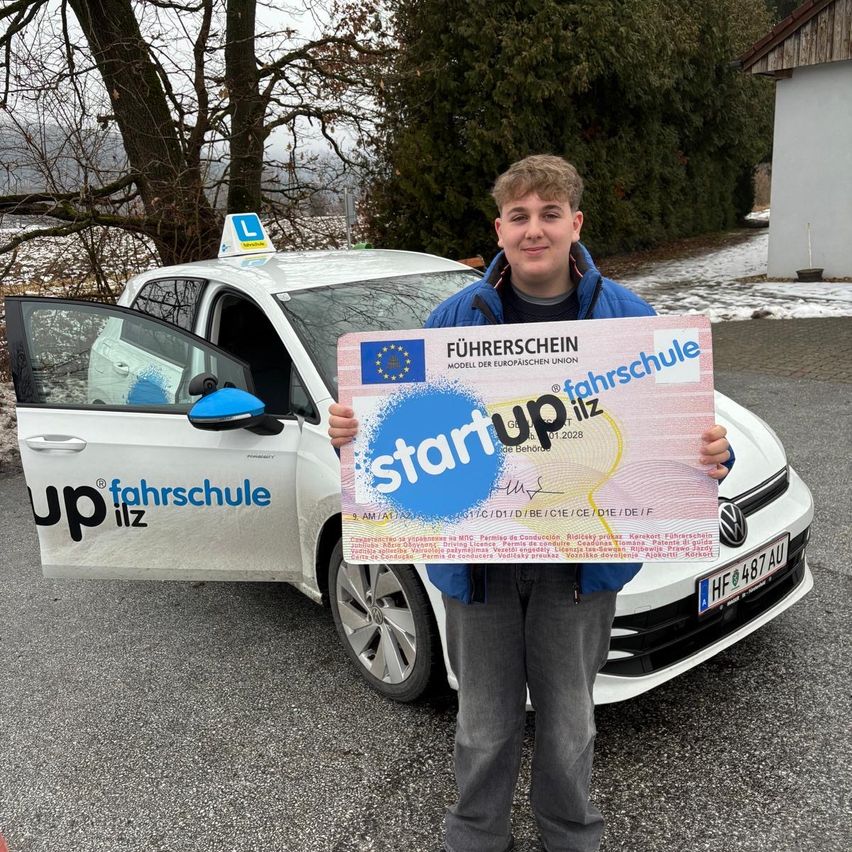 Ein junger Mann steht neben einem weißen Auto mit offener Tür und hält ein Zertifikat mit der Aufschrift 'Startup' und 'Fahrschule' auf Deutsch. Das Auto hat ein 'L' auf einem Schild über der Windschutzscheibe und ein Nummernschild mit der Aufschrift 'HF 487 AU'.