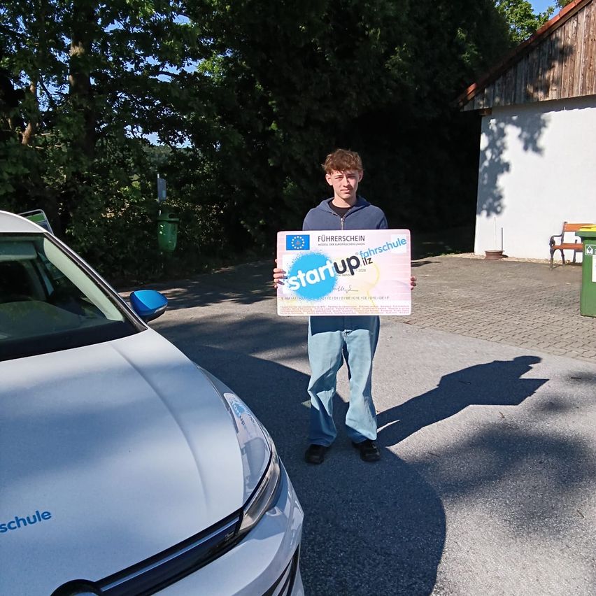 A man stands next to a white car, holding a certificate for a driving school.