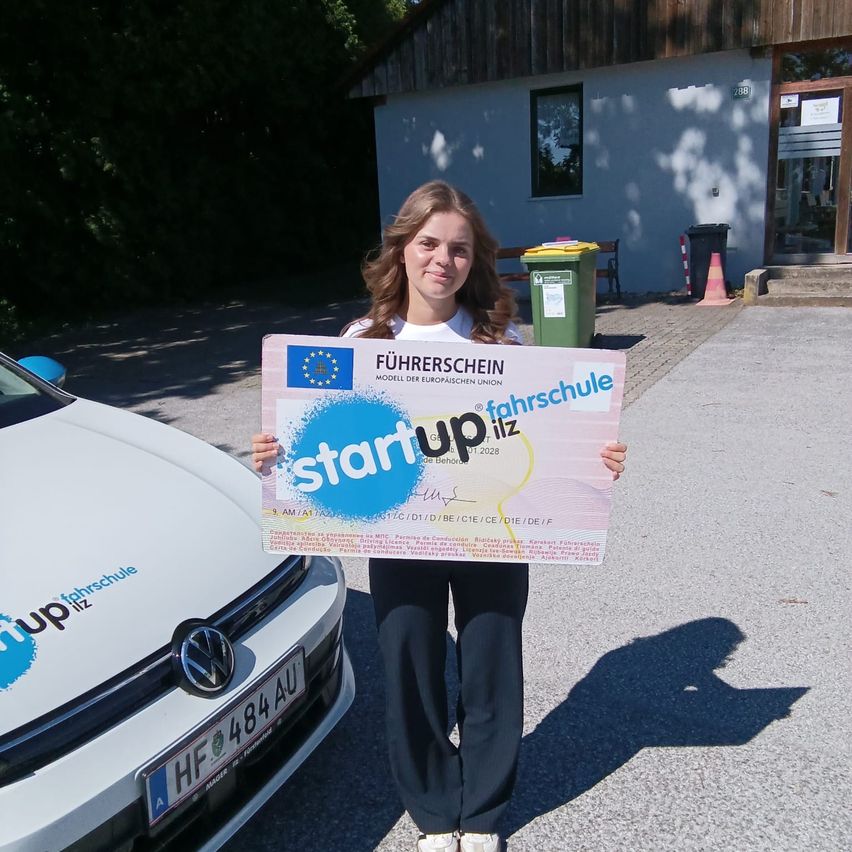 A woman holding a certificate from the European Union stands in front of a building with a car. The car's license plate reads 'HF 484 AU'.