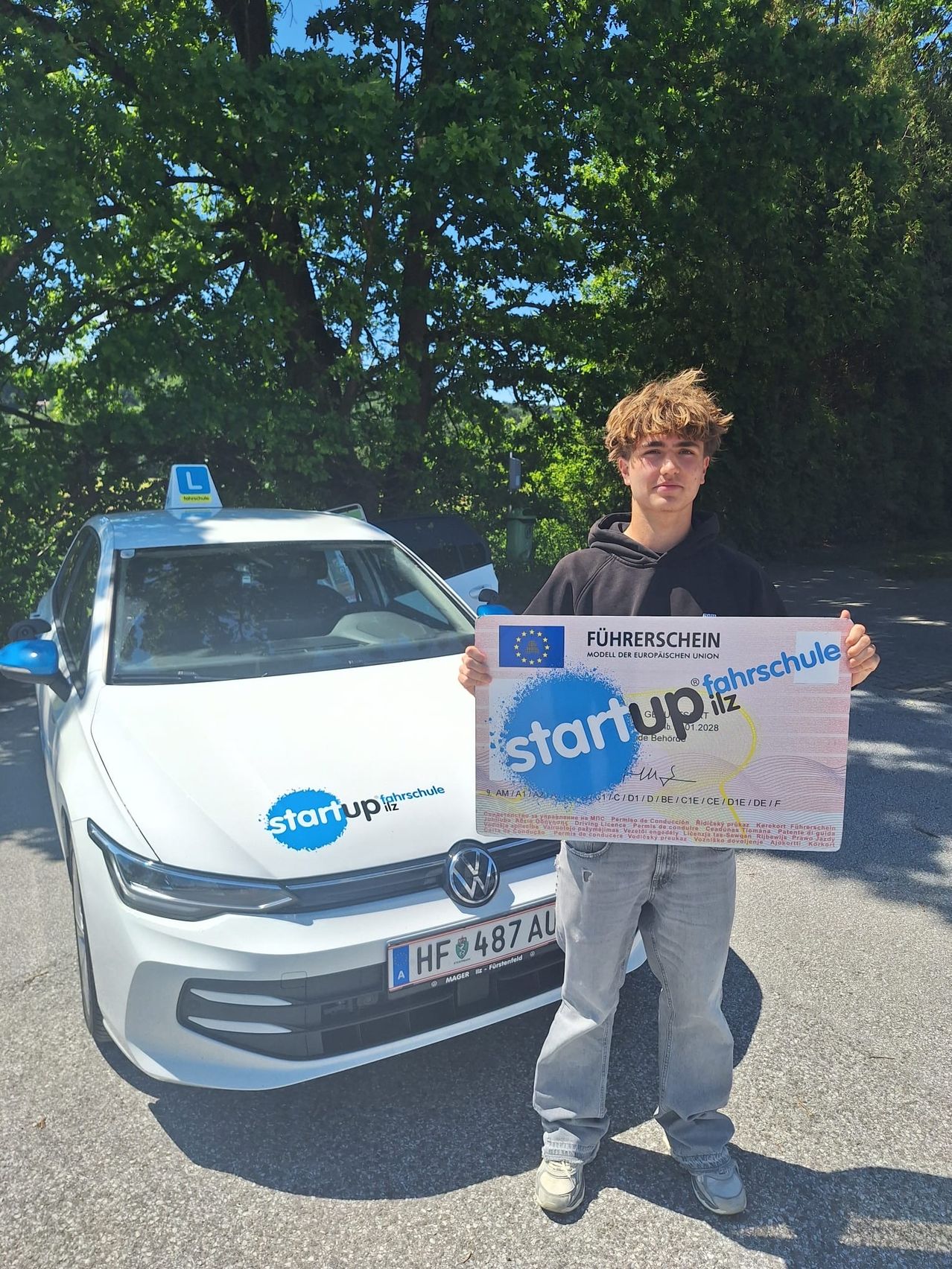 A young man stands outside a white car, holding a certificate for a driving school. The certificate is titled 'Start up Fahrschule'. The car has a taxi sign on top.