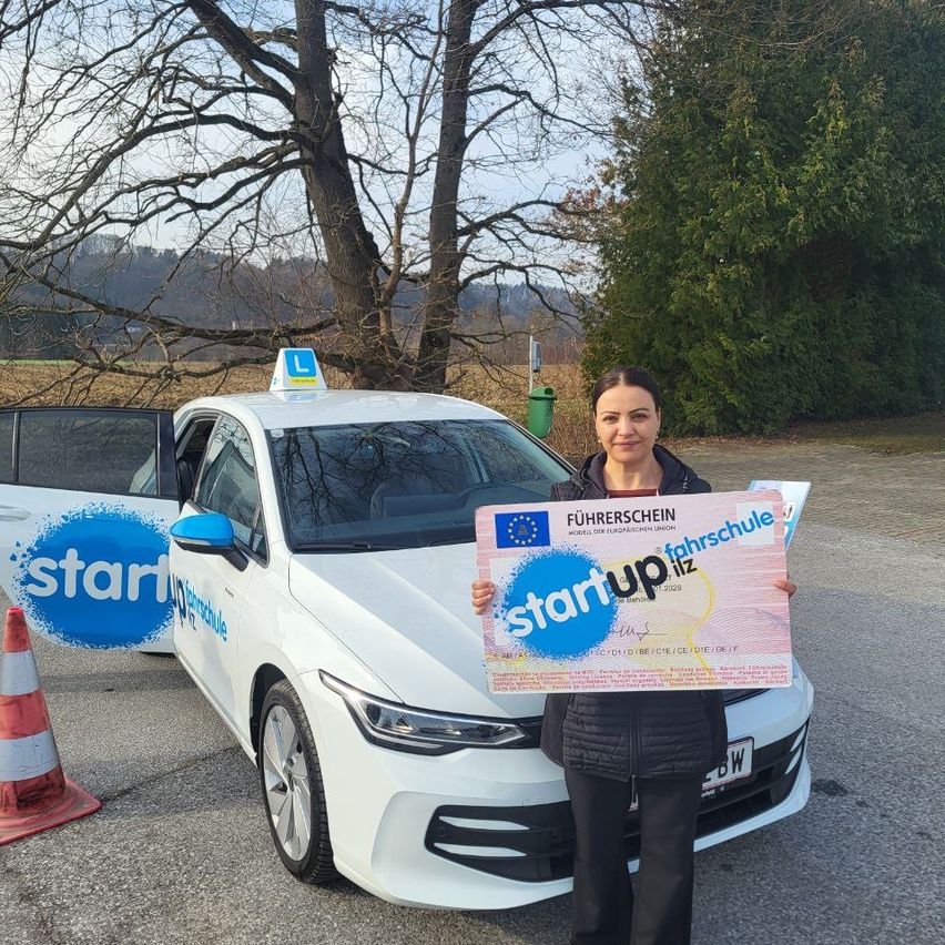 Eine Frau steht neben einem weißen Auto mit dem Wort 'start' an der Tür. Sie hält ein Zertifikat mit den Worten 'start up' und 'fahrschule'. Ein Verkehrskegel steht auf der linken Seite des Autos.