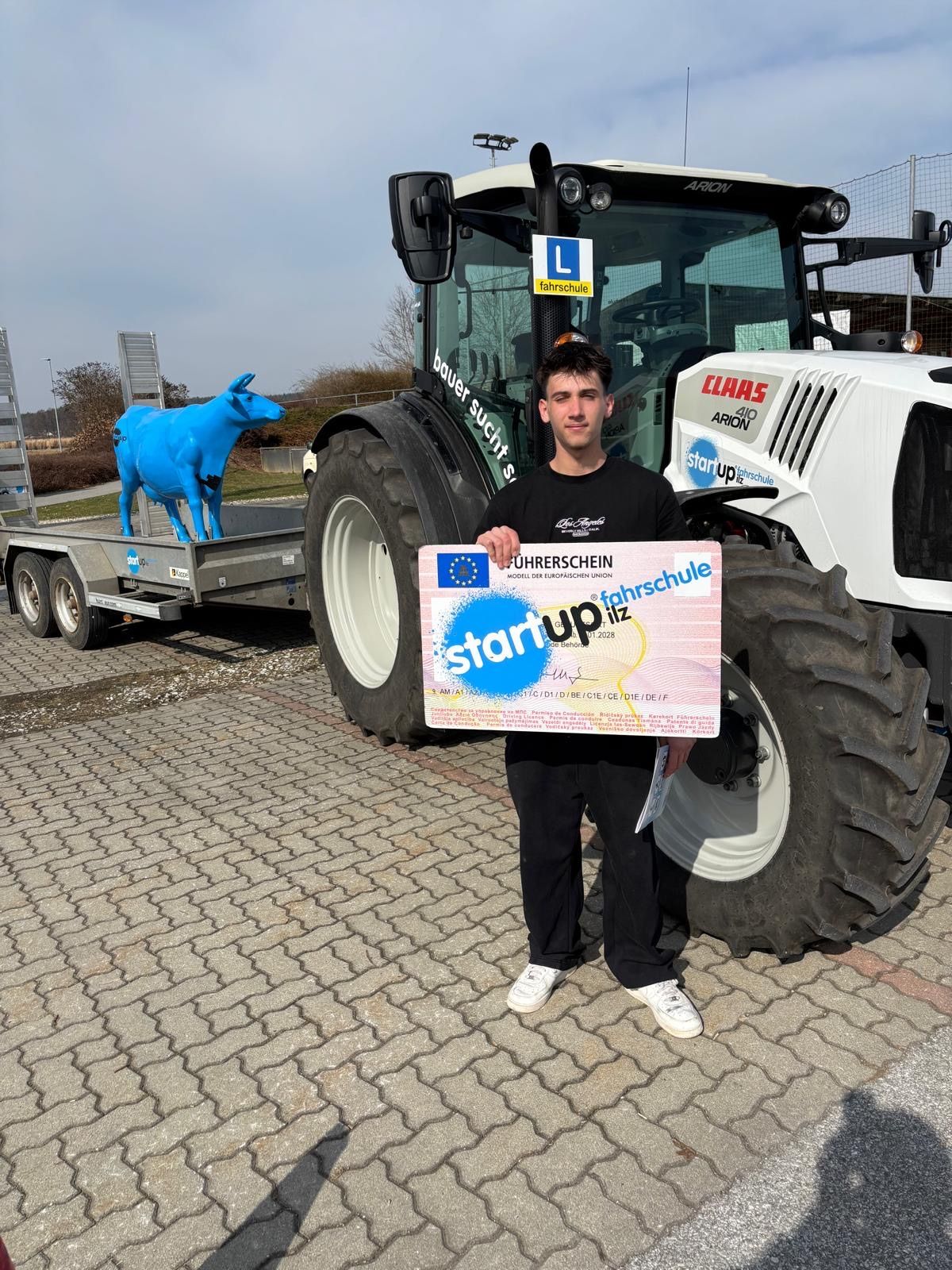 Ein junger Mann steht neben einem Traktor und hält ein Zertifikat in der Hand, im Hintergrund befindet sich eine Kuhstatue.