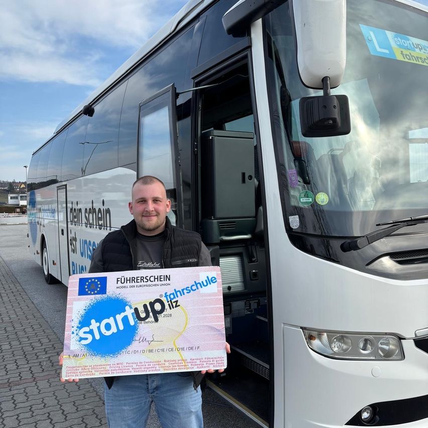 Ein Mann in einer schwarzen Weste hält einen rosafarbenen Gutschein mit der Aufschrift 'Start up', vor sich. Die Bustür ist hinter ihm geöffnet. Der Bus hat den Text 'FUHRERSCHEIN' an der Seite. Der Mann steht auf einem gepflasterten Weg.