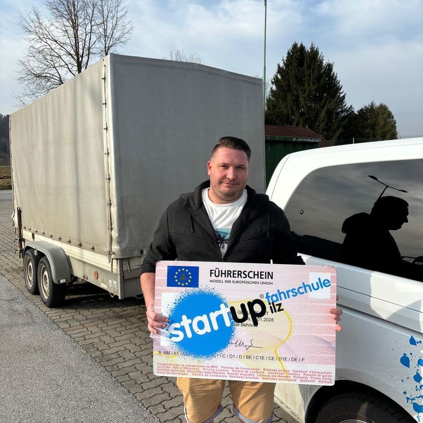 Ein Mann hält ein Zertifikat für Startup Fahrschule vor einem Fahrzeug und einem Anhänger auf einer Straße mit Bäumen im Hintergrund.