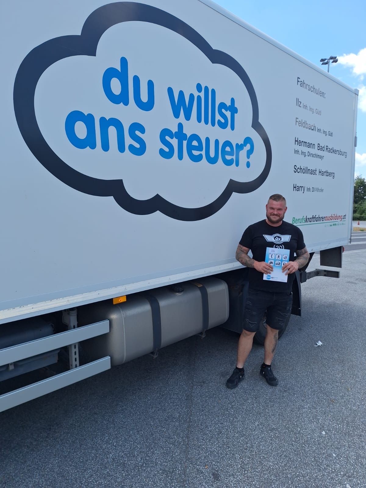 A man stands in front of a truck with a sign that reads 'Do you want to drive?' in German. He holds a certificate.