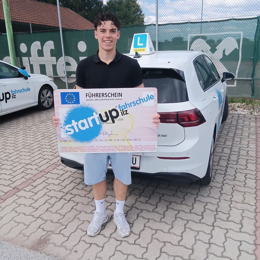 A young man stands in front of a white car, holding a certificate that reads 'Start up' and 'Führerschein'. He is wearing a black shirt and shorts. Behind him is a fence with logos and text.