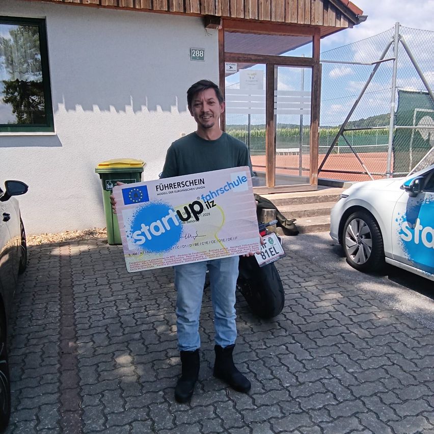 A man stands in front of a building holding a large sign that reads 'Start up' in blue and white. He is smiling and wearing a green shirt and jeans. Behind him, there is a trash bin and a car parked in front of a building with a glass door.