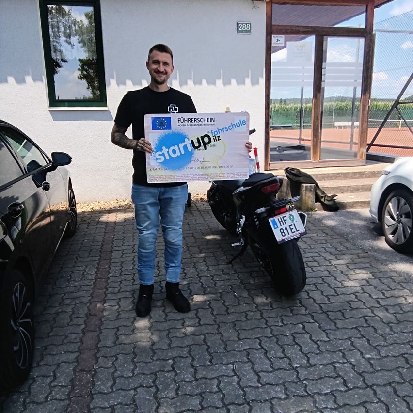 A man stands in front of a building holding a certificate labeled 'Startup Schule'. He is wearing a black shirt and jeans. Behind him, a motorcycle and two cars are parked on a brick pavement.