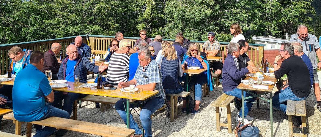 Eine Gruppe von Menschen sitzt an Tischen in einem Außenbereich, einige tragen Hüte, Brillen und Turnschuhe. Flaschen, Gläser und Teller stehen auf den Tischen. Der Bereich hat einen Zaun und Bäume im Hintergrund.