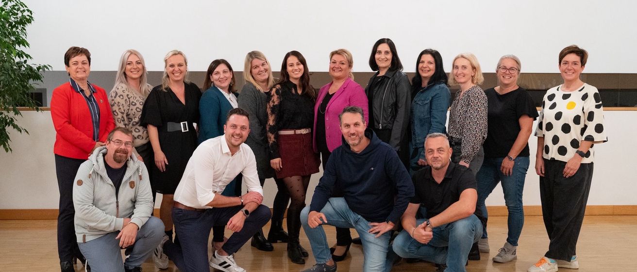 Eine Gruppe von Menschen, sowohl Männer als auch Frauen, posiert für ein Foto auf einem Holzboden. Sie alle lächeln und einige tragen Brillen. Sie sind in verschiedenen Outfits gekleidet. Die Wand hinter ihnen ist weiß.