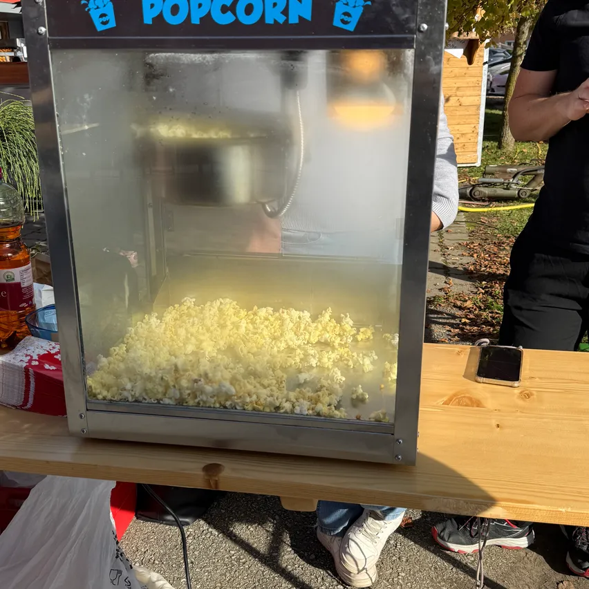 Eine Popcornmaschine steht auf einem Holztisch mit einer Hand darin. Die Maschine ist mit Popcorn gefüllt. Ein Smartphone liegt auf dem Tisch.