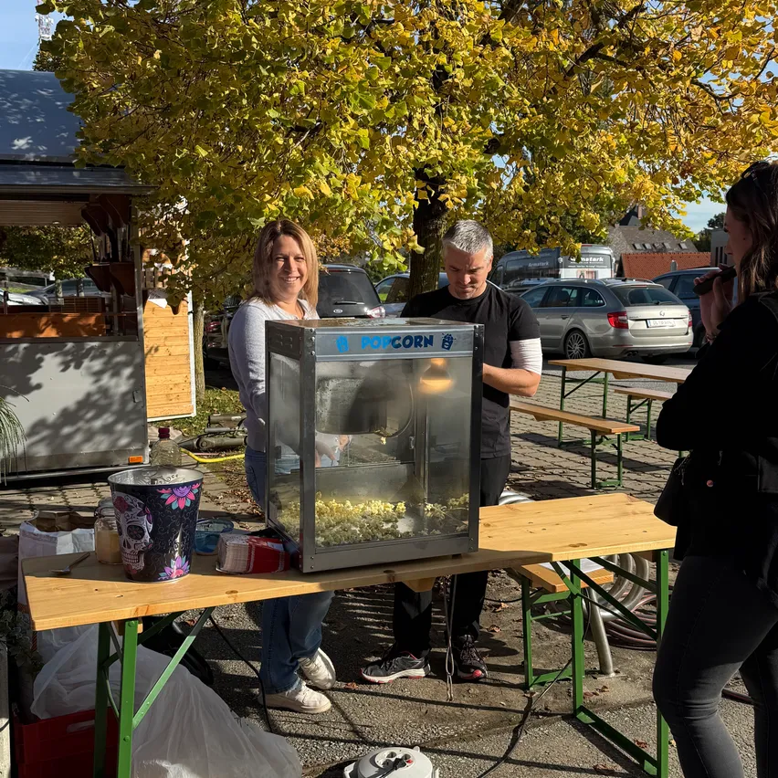 Zwei Personen stehen neben einer Popcornmaschine auf einem Tisch im Freien. Eine Frau lächelt und ein Mann schaut sie an. Eine Frau rechts macht ein Foto.