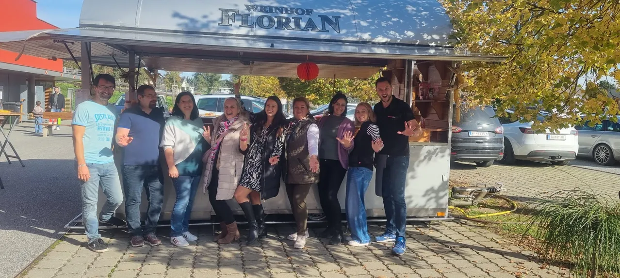 Eine Gruppe von Frauen und einem Mann posiert für ein Foto vor einem Florian Weinladen an einem sonnigen Tag.