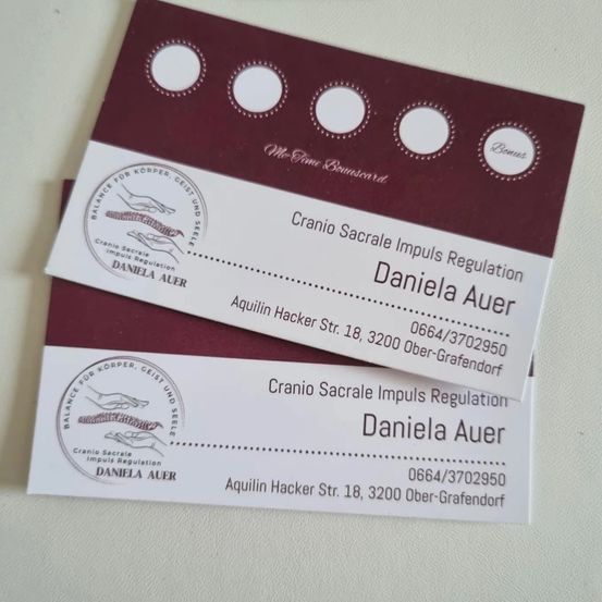 Zwei Visitenkarten sind auf einer weißen Oberfläche zu sehen. Die Karten haben einen kastanienbraunen Hintergrund und weißen Text. Die Karten haben ein Logo und Text, der 'Cranio Sacrale Impuls Regulation Daniela Auer' lautet. Die Karten haben auch eine Telefonnummer und Adresse. Die Karten haben einen Bonusaufkleber oben rechts.
