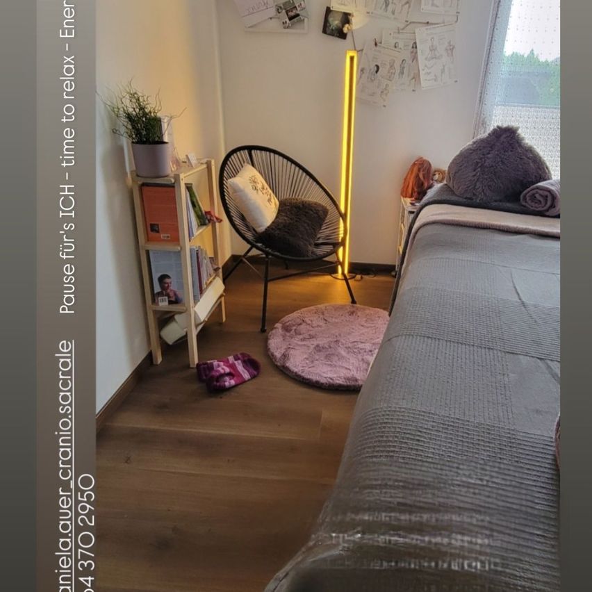 A cozy bedroom with a bed, a round rug, and a chair. A wooden shelf holds a potted plant, books, and a photo. A yellow lamp and some papers are on the wall.
