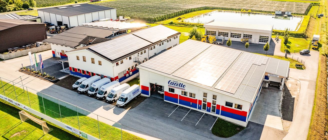 Luftaufnahme einer Fabrik mit mehreren Gebäuden und Lieferwagen. Solarpanele bedecken die Dächer. Grüne Felder und Bäume im Hintergrund.