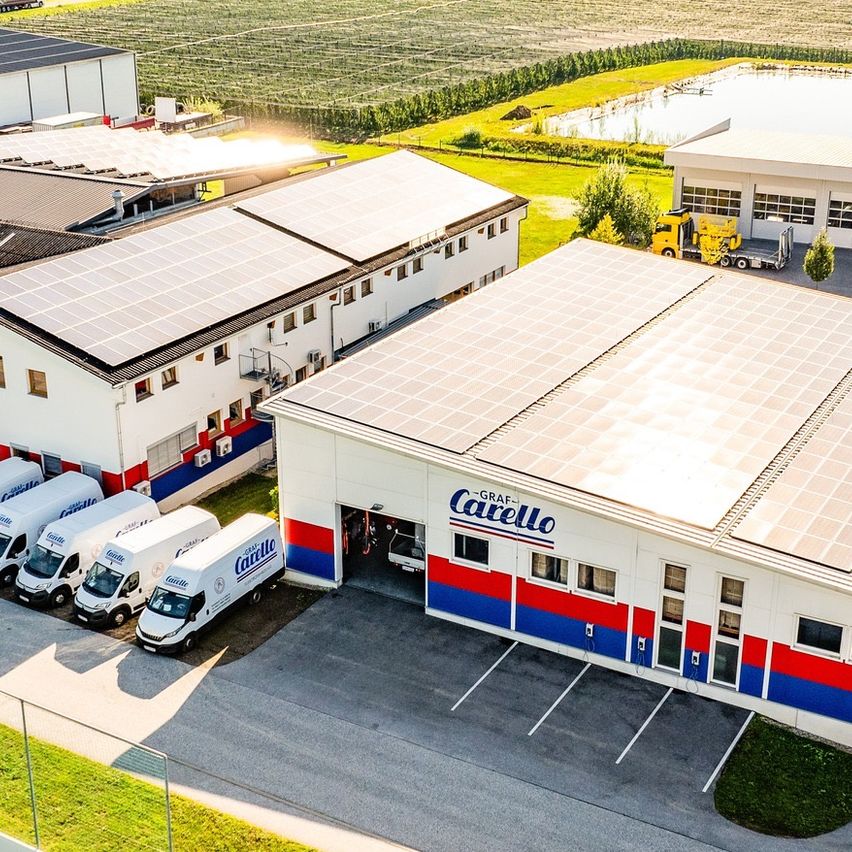 Luftaufnahme einer Fabrik mit mehreren Gebäuden und Lieferwagen. Solarpanele bedecken die Dächer. Grüne Felder und Bäume im Hintergrund.