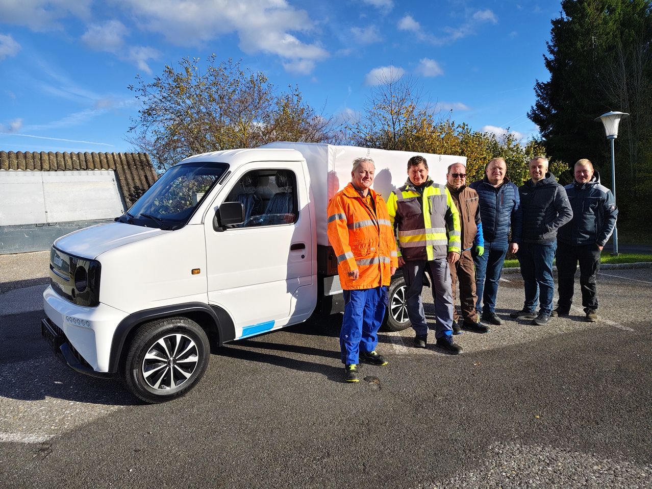 Fünf Männer stehen neben einem weißen Lieferwagen unter einem blauen Himmel mit Bäumen im Hintergrund.