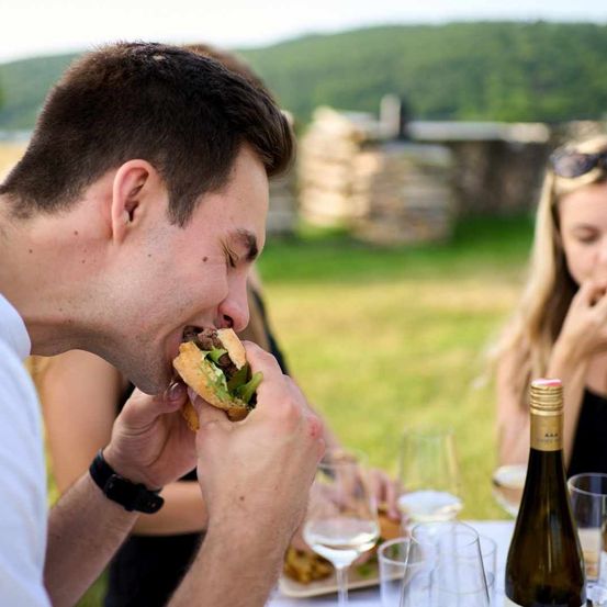 Ein Mann genießt im Freien einen Sandwich mit Freunden in der Nähe einer Steinmauer. Weingläser und eine Flasche Wein stehen auf dem Tisch.