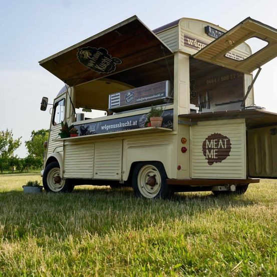 Ein Food-Truck mit offenem Vordach, der ein Markenlogo und den Slogan 'Meat Me' zeigt. Er steht auf einem Rasenfeld mit Bäumen im Hintergrund.