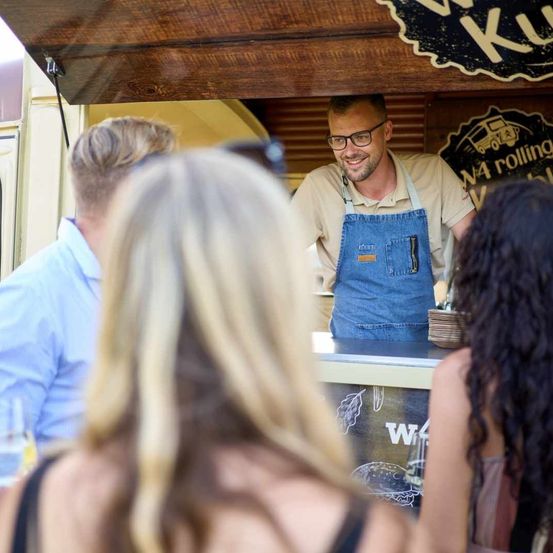 Ein Mann in einer Schürze steht lächelnd hinter einem Tresen in einem Food Truck. Vor ihm stehen drei Personen, zwei Frauen und ein Mann.