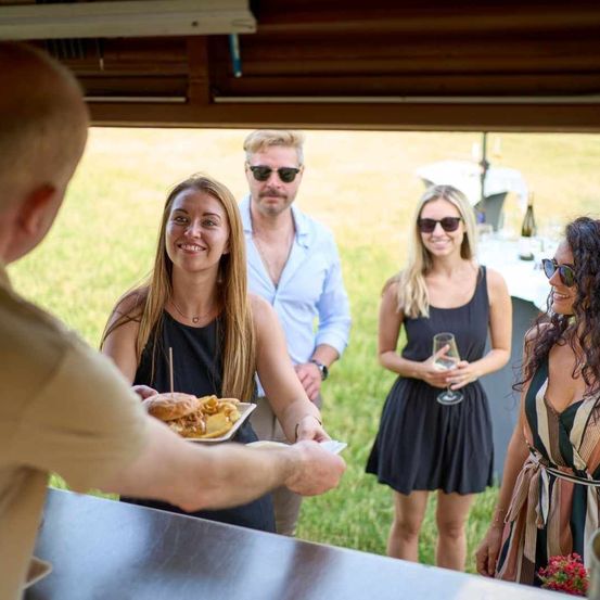 Ein Mann serviert Essen auf einem Teller einer Frau, die in der Nähe eines Tisches steht. Dahinter stehen ein Mann und eine Frau mit Sonnenbrillen und Kleidern in der Nähe eines Tisches mit Weingläsern.