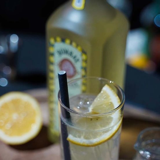 Ein Glas Zitronengetränk mit Strohhalm, einer Zitronenscheibe und einer Ginflasche im Hintergrund.